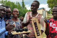A group of smiling individuals outdoors holding handmade tools and materials, showcasing a participatory design project.